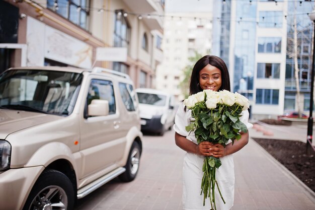 Schönes afrikanisch-amerikanisches Mädchen, das einen Strauß weißer Rosen hält, blüht bei der Datierung in der Stadt Schwarze Geschäftsfrau mit Blumenstrauß in der Nähe des Autos