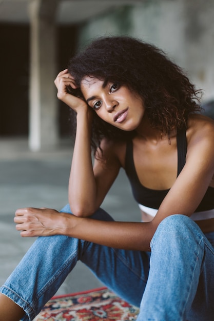 Schönes Afroamerikanermädchen mit dunklem lockigem Haar im schwarzen sportlichen Oberteil und in den Jeans nachdenklich beim Sitzen auf Vintage-Teppich zu Hause