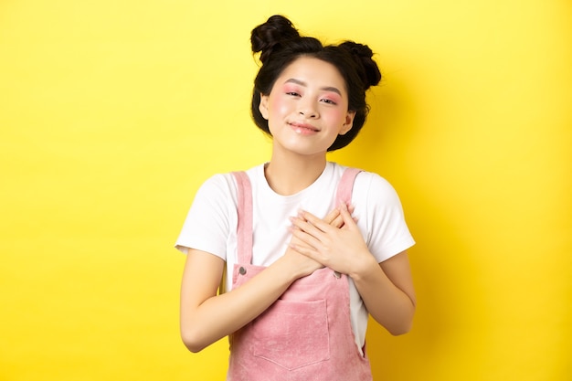 Schönes asiatisches jugendlich Mädchen in der Liebe, Händchen haltend am Herzen und lächelnd berührt, mit Zuneigung schauend, auf gelb stehend.