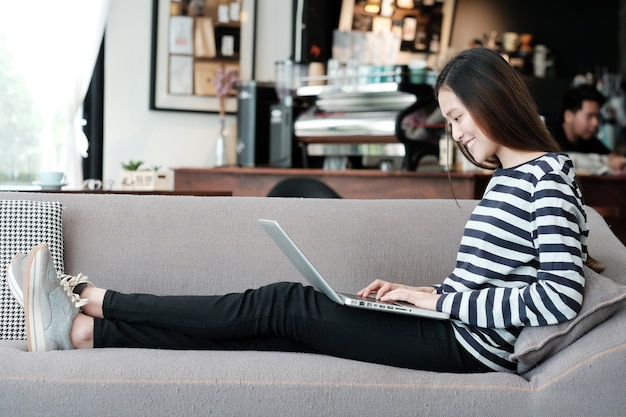 Schönes asiatisches Mädchen, das Laptop-Computer beim Sitzen auf Sofa mit lächelndem Gesichtsgefühl verwendet