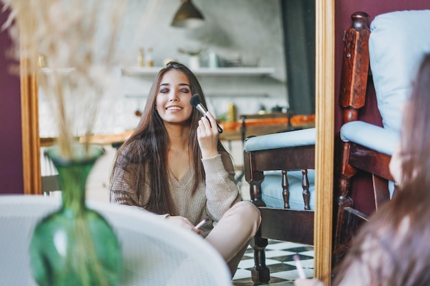 Schönes asiatisches Mädchen mit langen Haaren, das sich den Spiegel anschaut und Make-up mit dem Pinsel macht, der zu Hause auf dem Boden sitzt