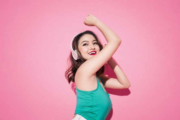 Schönes asiatisches Mädchen mit professionellem Make-up und stilvoller Frisur, das singt und tanzt, während es Musik auf rosafarbenem Hintergrund hört.