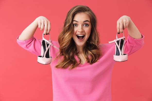 Schönes aufgeregtes junges Mädchen in Freizeitkleidung, das isoliert über rosa Wand steht und ein Paar Schuhe mit hohen Absätzen zeigt