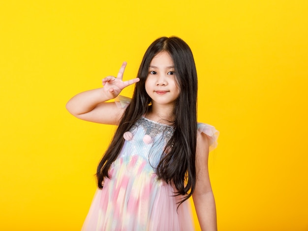 Schönes ausgeschnittenes Porträt eines jungen hübschen asiatischen Mädchens auf einem süßen, lässigen Kinderkleid macht eine attraktive Fingergeste als selbstbewusstes Siegeszeichen mit fröhlichem Gesicht, um den sicheren Erfolg auszudrücken, Garantie erfüllt