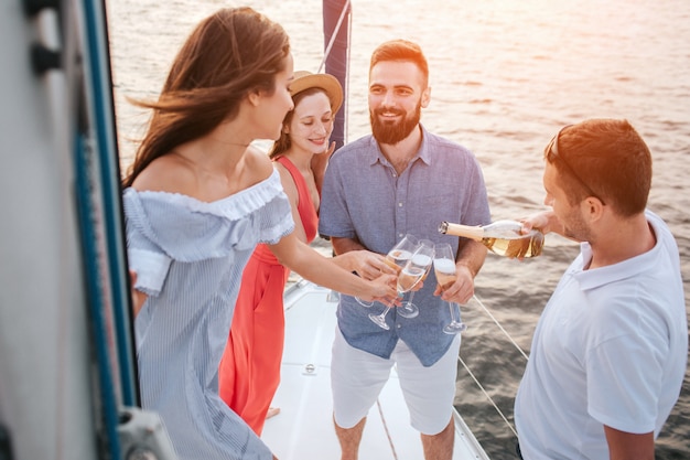 Schönes Bild von vier Personen, die zusammen auf der Yacht stehen. Mann im weißen Hemd gießt Champagner in Gläser. Brünette sieht einen anderen Mann an.