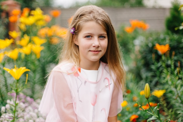 schönes blauäugiges Mädchen mit langen blonden Haaren. kleines Mädchen in einem rosa Flamingokleid. Mädchen im Blumengarten. Sommer helles, emotionales Foto. großer, dicker, heller Blumengarten in der Nähe des Hauses.