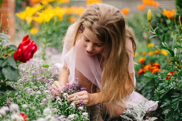 schönes blauäugiges Mädchen mit langen blonden Haaren. kleines Mädchen in einem rosa Flamingokleid. Mädchen im Blumengarten. Sommer helles, emotionales Foto. großer, dicker, heller Blumengarten in der Nähe des Hauses.