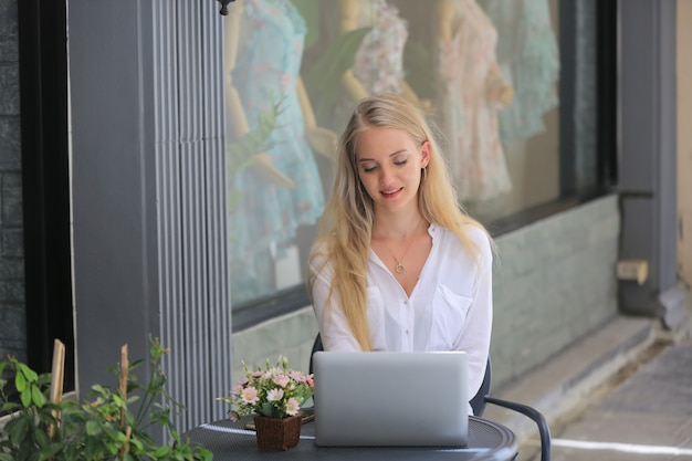 Schönes blondes Haarmädchen, das mit Laptop vor Einzelhandelsgeschäft sitzt, Kleinunternehmerkonzept