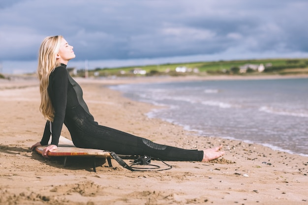 Schönes blondes im Taucheranzug mit Surfbrett am Strand