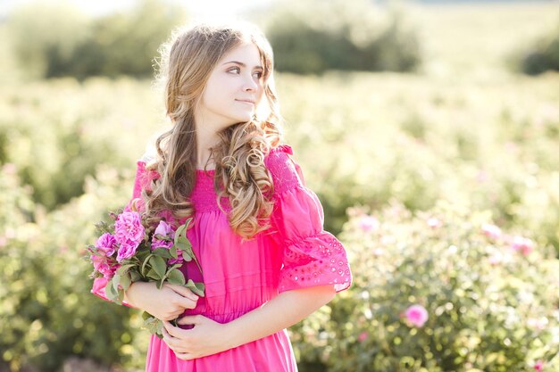 Foto schönes blondes jugendlich mädchen, das rosafarbenes kleid trägt, das draußen rosenblume hält