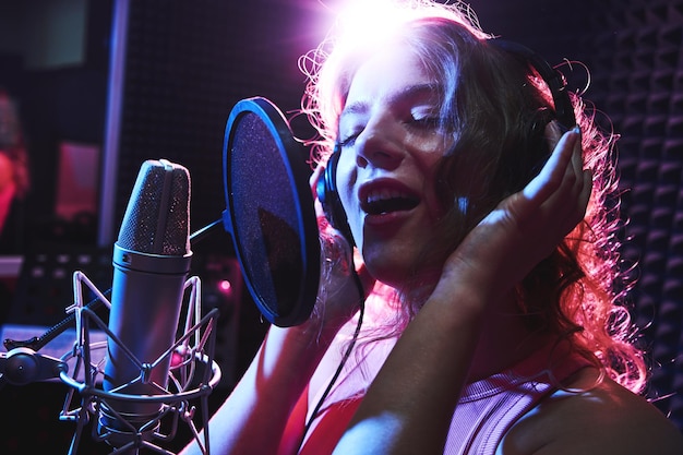 Schönes blondes Mädchen, das emotional ein Lied im Tonstudio mit professionellem Mikrofon und Kopfhörern singt, erstellt einen neuen Track-Album-Vokalkünstler in rosa-blauem Neonlicht-Nahaufnahme-Gesicht
