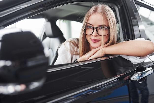 Schönes blondes Mädchen, das im neuen Auto mit modernem schwarzem Innenraum sitzt