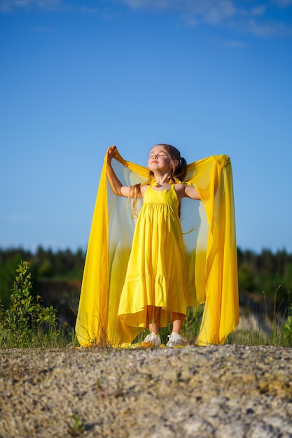 Schönes blondes Mädchen, das in einem gelben Kleid in der Natur aufwirft. Sommerfoto. Blauer Himmel. Sonniger Tag.