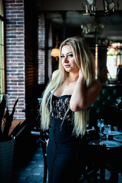 schönes blondes Mädchen, das in einem Restaurant in einem schwarzen Kleid steht und aufwirft