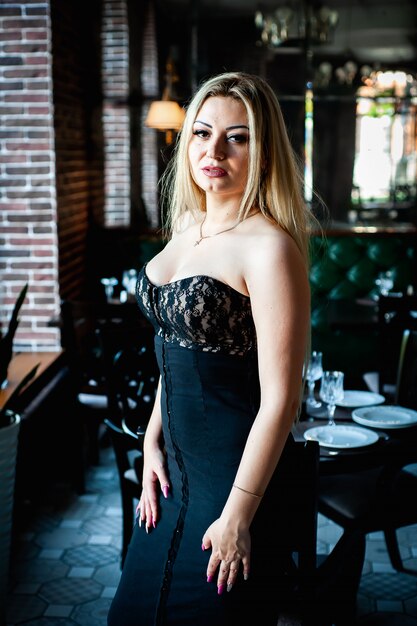 schönes blondes Mädchen, das in einem Restaurant in einem schwarzen Kleid steht und aufwirft