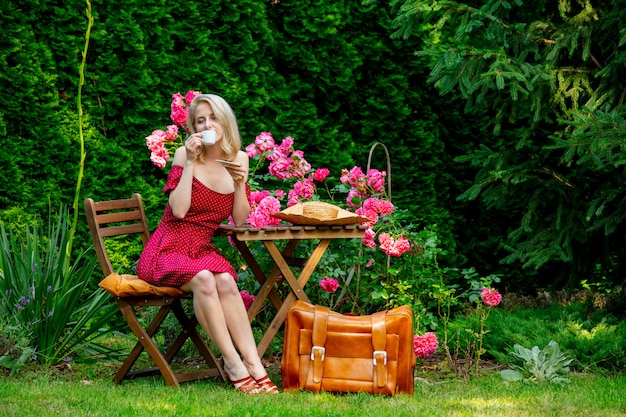 Schönes blondes Mädchen im roten Kleid mit Koffer trinkt einen Kaffee in einem Garten