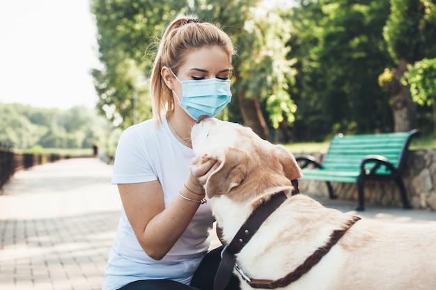 Schönes blondes Mädchen und ihr Labrador umarmen sich in einem Park, während sie eine medizinische Maske tragen