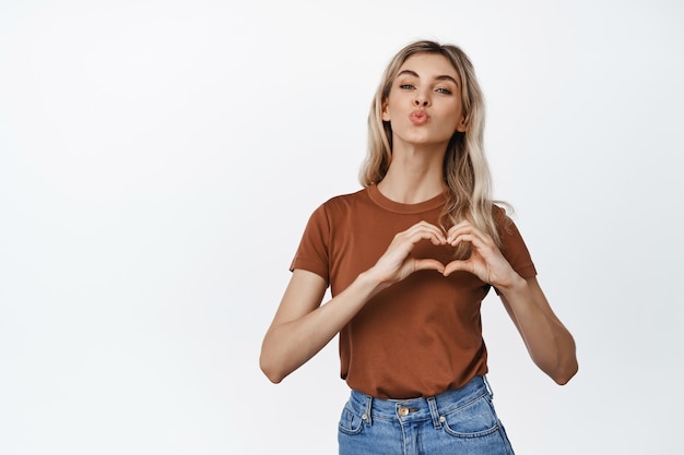 Schönes blondes Mädchen verzieht die Lippen im Kuss, zeigt Herzliebesgeste auf der Brust und steht auf Weiß.