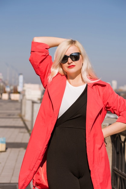Schönes blondes Model, das in rotem Umhang posiert, trägt eine Sonnenbrille auf einem verschwommenen Stadthintergrund
