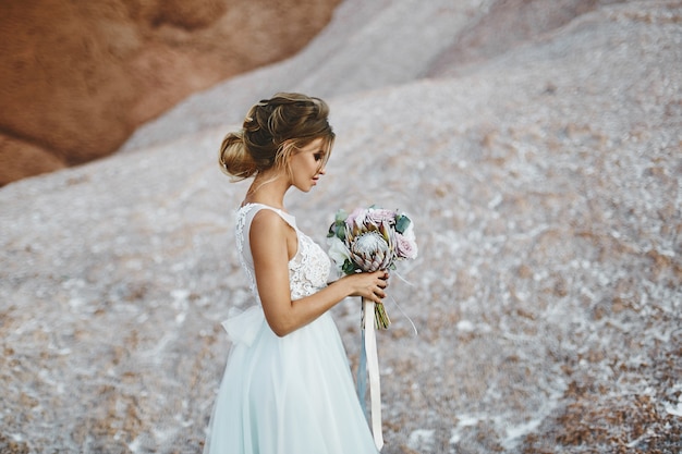 Schönes blondes Modellmädchen mit modellierender Hochzeitsfrisur in einem modischen weißen Spitzenkleid mit einem Blumenstrauß von exotischen Blumen in ihren Händen