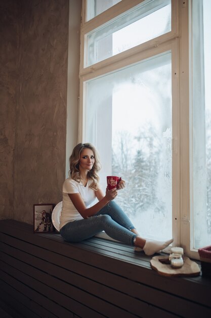 Schönes blondes Sitzen auf Fensterbrett mit Becher