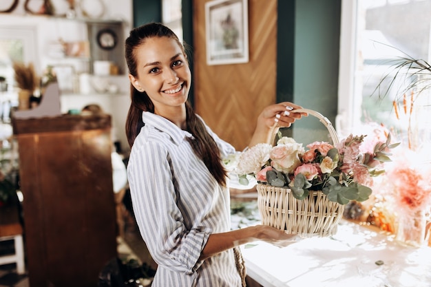 Schönes brünettes Mädchen, das in einem gestreiften Kleid gekleidet ist, hält Blumenstrauß aus frischen Blumen in Pastellfarben im Korb im Blumenladen.