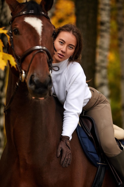 Schönes brünettes Mädchen der Herbstlandschaft mit dem langen Haar, das mit einem roten Pferd im Wald aufwirft