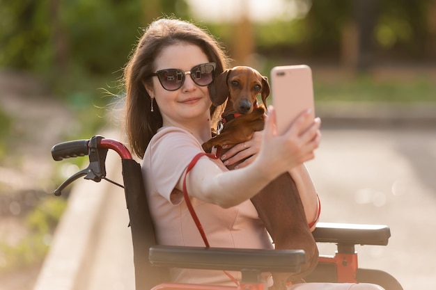 schönes brünettes mädchen im rollstuhl im sommer auf einem spaziergang mit einem süßen dachshund hund nimmt ein se