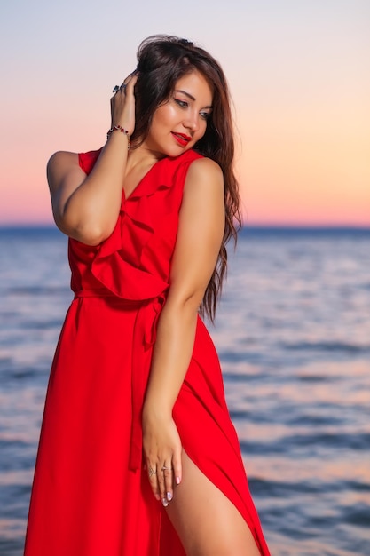 Schönes brünettes Mädchen in einem roten Kleid, das bei Sonnenaufgang oder Sonnenuntergang an der Küste posiert