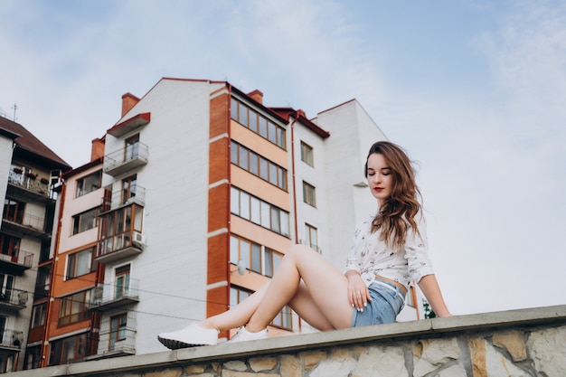 Schönes brünettes Mädchen in T-Shirt und Shorts sitzt auf einem Hintergrund großer Häuser. Mädchen mit langen Haaren posiert auf dem Hintergrund der Sommerstadt.