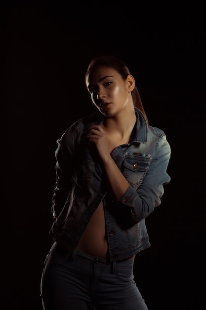 Schönes brünettes Model in trendiger Jeanskleidung posiert im dunklen Studio