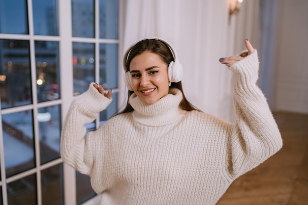 Schönes brünettes weibliches Model in weißem Pullover und Kopfhörern, das nach Hause gegen das Fenster tanzt