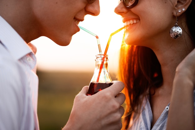 Schönes dunkelhaariges Mädchen und ein junger Mann mit Sonnenbrille trinken an einem sonnigen Tag aus einer Flasche ein Getränk durch Strohhalme im Freien. .