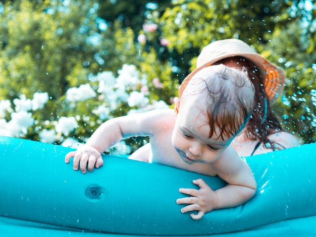 Schönes einjähriges Mädchen badet und spielt an einem heißen Tag in einem aufblasbaren Pool mit Mama