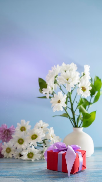 Schönes festliches Stillleben mit einem Geschenk in roten Papierweißblumen in weißer Vase auf blauem Hintergrund