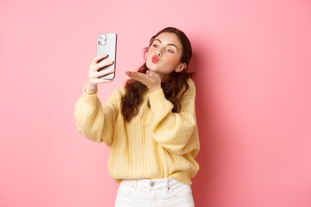 Schönes flirty Mädchen, das selfie auf Smartphone nimmt, Luftkuss während des Video-Chats sendet, der gegen rosa Wand steht. Speicherplatz kopieren