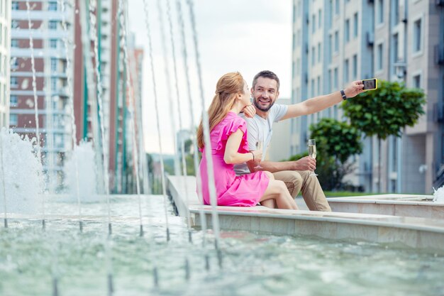 Schönes Foto. Netter positiver Mann, der zusammen mit seiner Freundin sitzt und ein Selfie mit ihr macht