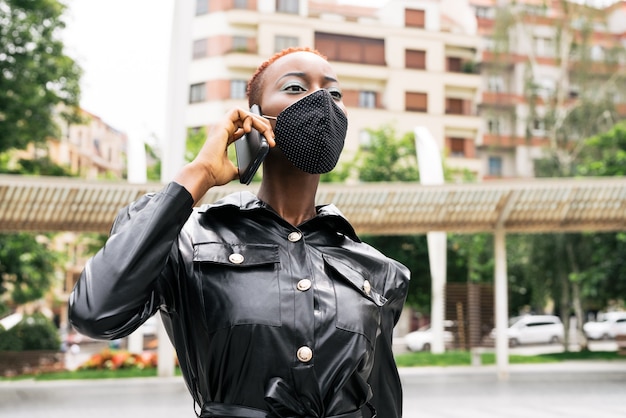 Schönes Frauenmodell mit Maske wegen der Coronavirus-Pandemie covid 19, die auf ihrem Handy spricht und sich um ihr Geschäft auf der Straße kümmert