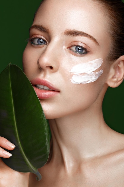 Schönes frisches Mädchen mit kosmetischer Creme auf dem natürlichen Make-up des Gesichts und grünen Blättern Schönheitsgesicht