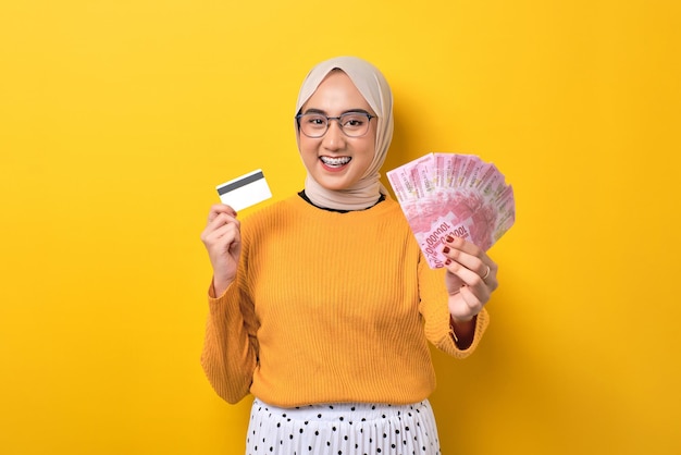 Schönes fröhliches asiatisches Mädchen mit Hijab, das Geldbanknoten und Kreditkarte isoliert auf gelbem Hintergrund zeigt