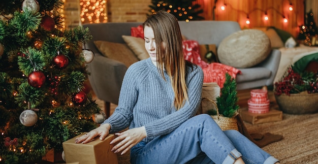 Schönes fröhliches glückliches junges Mädchen legt Weihnachtsgeschenke auf dem Boden nahe dem Neujahrsbaum zu Hause nieder