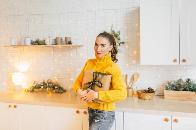 Schönes fröhliches glückliches junges Mädchen mit Weihnachtsgeschenken auf dem Hintergrund der Neujahrsküche