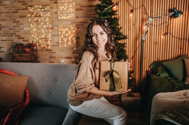 Schönes fröhliches glückliches junges Mädchen mit Weihnachtsgeschenken auf Sofa auf dem Hintergrund eines Neujahrsbaums zu Hause