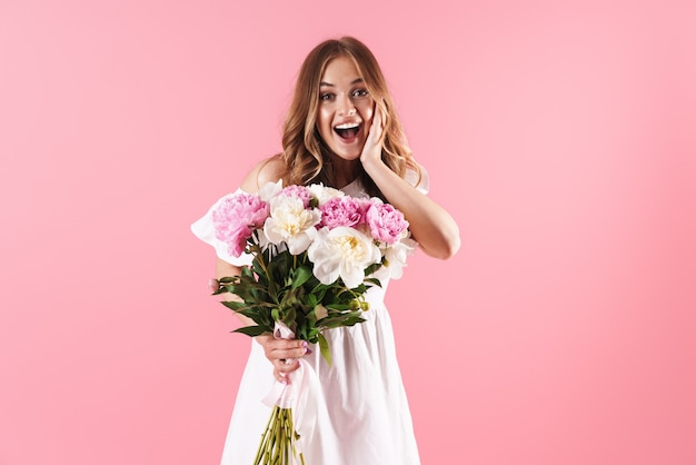 Schönes fröhliches junges blondes Mädchen, das Sommerkleid trägt, das isoliert über rosa Wand steht und einen Strauß Pfingstrosen hält