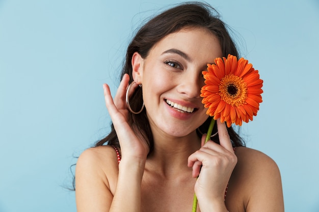 Schönes fröhliches Mädchen, das Sommerkleidung trägt, die lokal über Blau steht und mit einer Herbera-Blume aufwirft