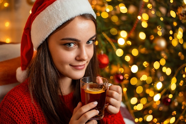 Schönes fröhliches Mädchen im Weihnachtshut, das Tasse Tee mit Zitronenscheibe hält