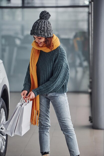 Schönes fröhliches Mädchen in gelbem Schal und in warmer Kleidung, die drinnen mit Einkaufstüten in den Händen steht.