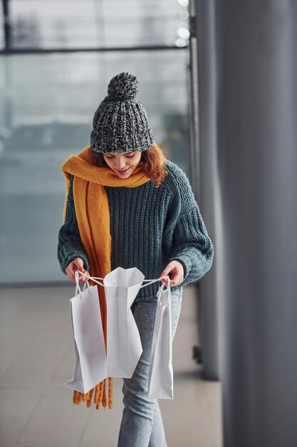 Schönes fröhliches Mädchen in gelbem Schal und in warmer Kleidung, die drinnen mit Einkaufstüten in den Händen steht.