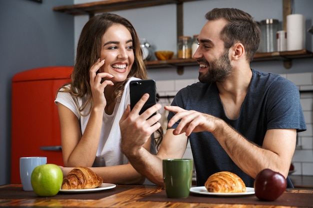 Schönes fröhliches Paar beim Frühstück in der Küche, mit Handy