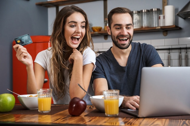 Schönes fröhliches Paar beim Frühstück in der Küche, mit Laptop-Computer, Kreditkarte zeigend